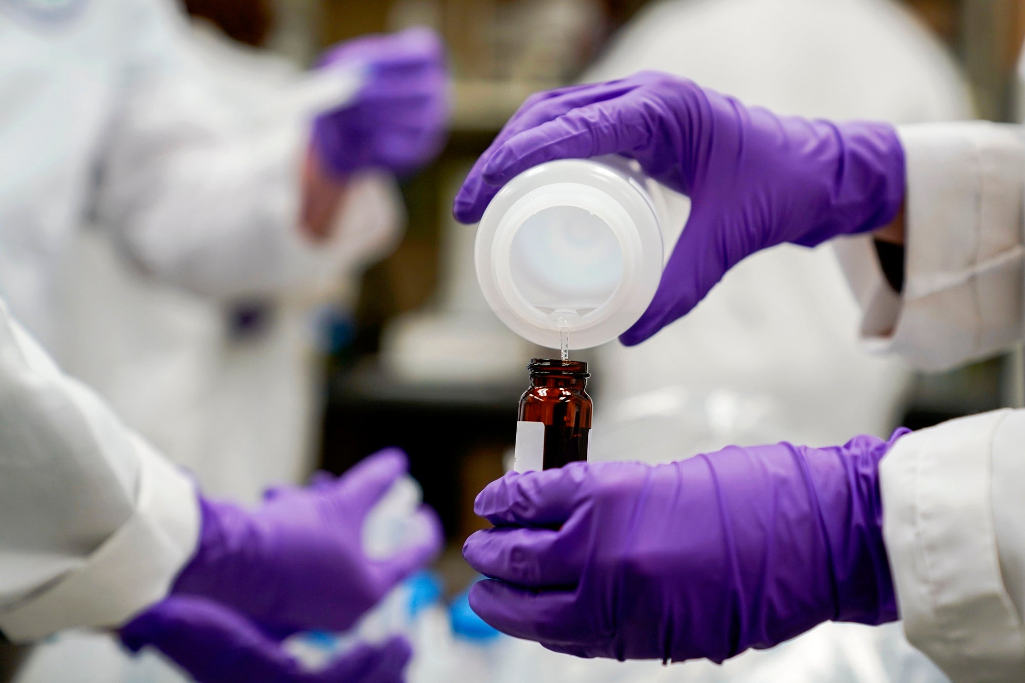 PFAS Pollution A close up of rubber gloved hands pouring water from one container to another in a laboratory.